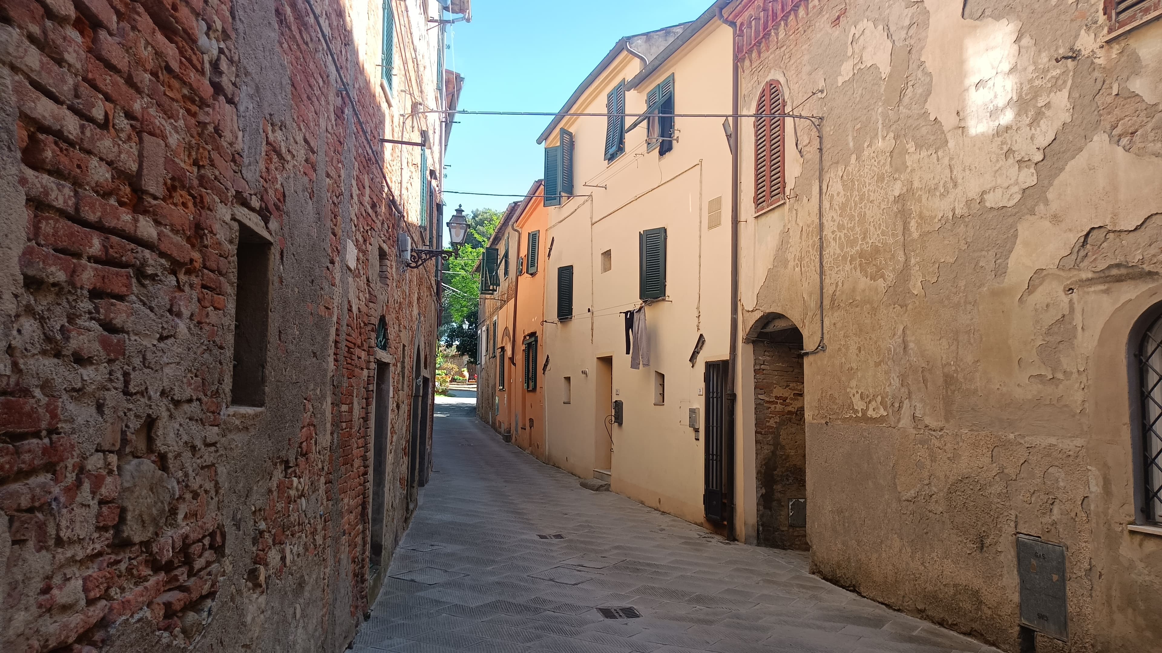 Narrow cobblestone alleyway between old brick and cream buildings with green shutters under blue sky.