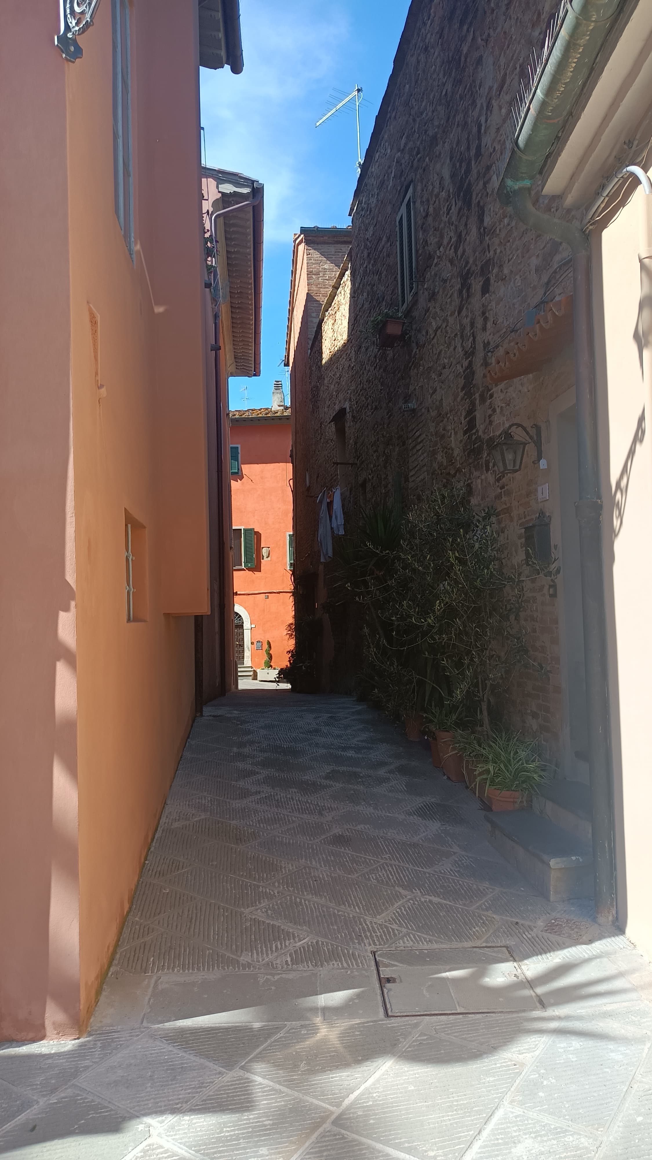Narrow paved alleyway between a peach building and a rustic stone wall with plants.