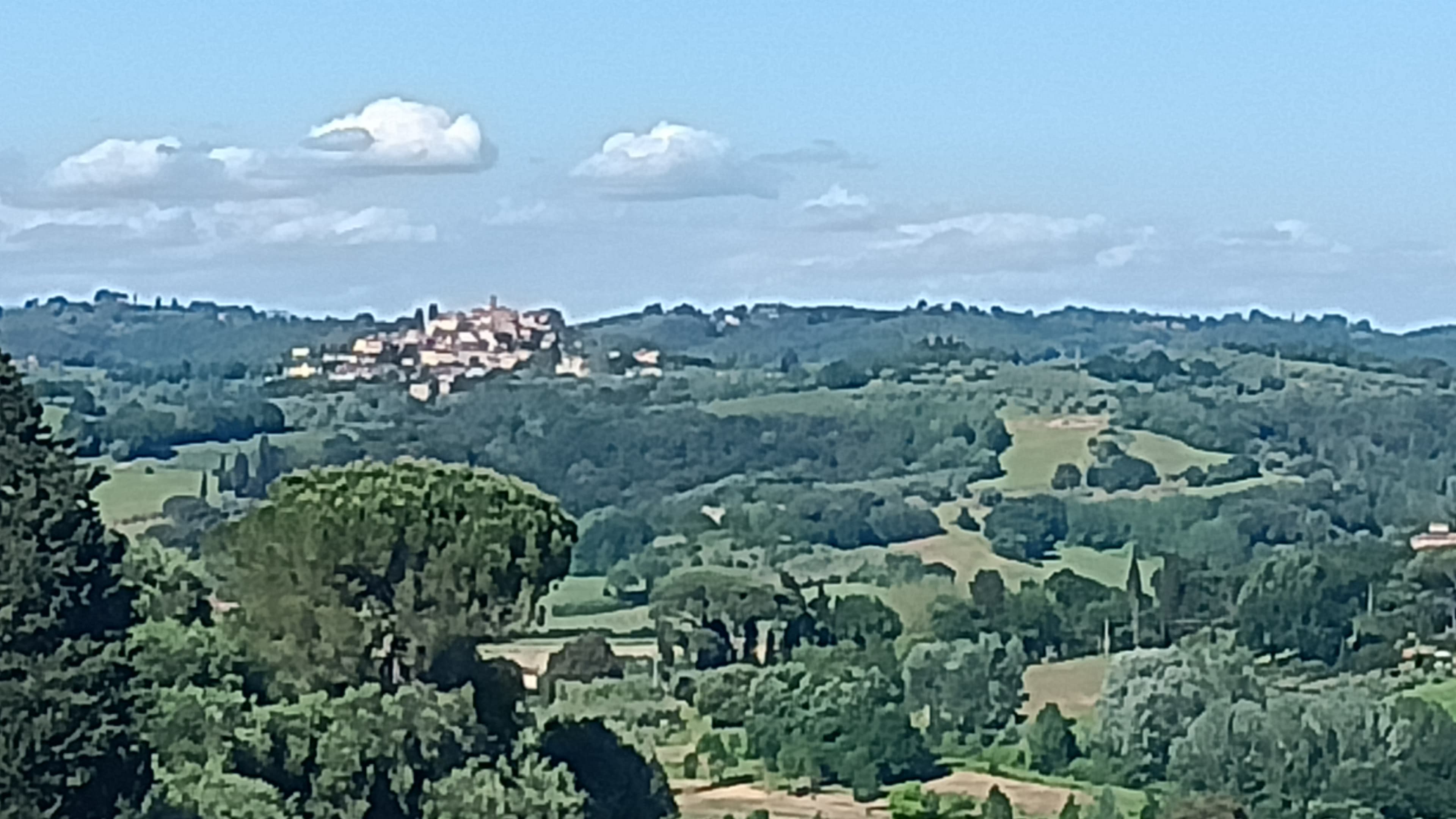 Lush green rolling hills with a distant hilltop village under a blue, cloudy sky.